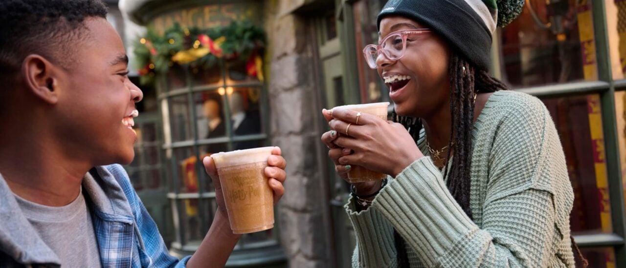 Ush Christmas 2026 Butterbeer Hogsmeade | 2026 好萊塢環球影城聖誕節&跨年派對攻略|Grinchmas 聖誕樹點燈+霍格華茲城堡聖誕光雕秀 兩位遊客在掛上聖誕裝飾的活米村街上,拿著奶油啤酒說笑