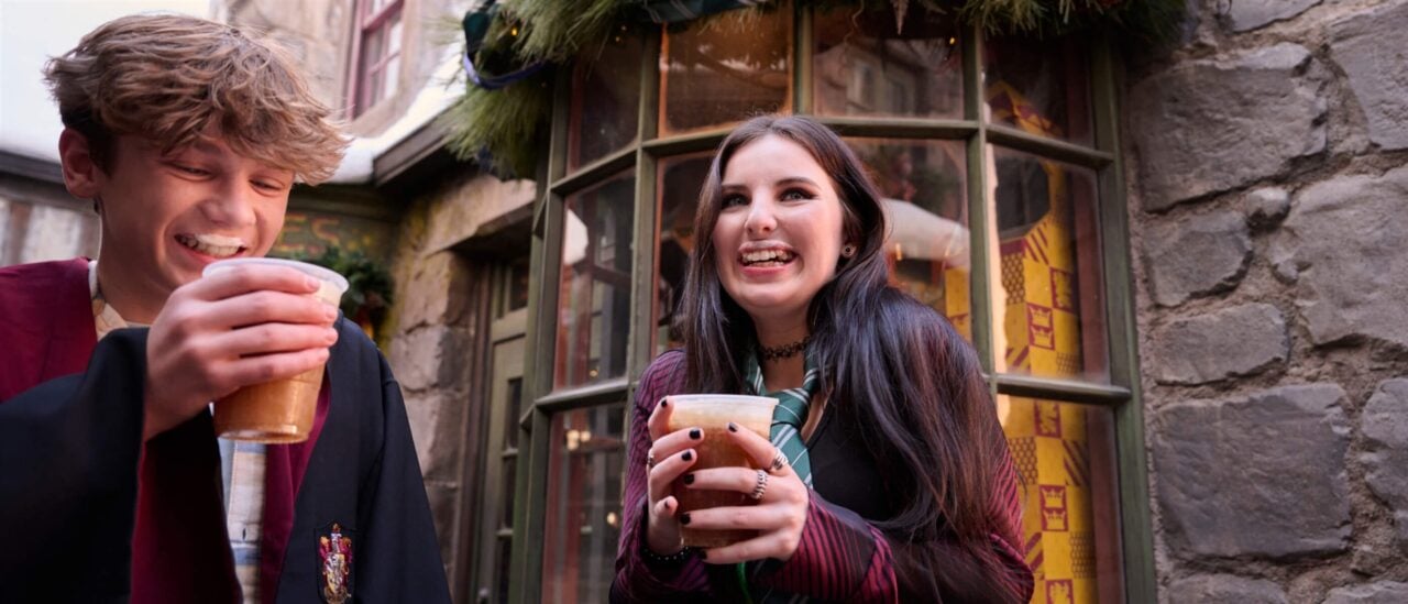 Ush Christmas 2026 Butterbeer Students | 2026 好萊塢環球影城聖誕節&跨年派對攻略|Grinchmas 聖誕樹點燈+霍格華茲城堡聖誕光雕秀 穿著霍格華茲學院袍的兩位遊客,在掛著聖誕花環的店門前喝奶油啤酒